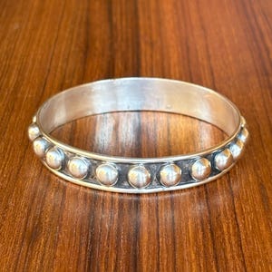 May include: A silver bangle bracelet featuring a continuous row of polished, raised spherical studs along the center. The metal has a smooth, reflective finish with darkened recessed areas between the circular elements, creating a distinct contrast.