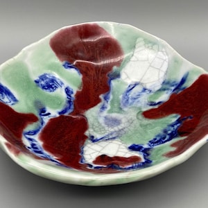 May include: A small, white ceramic bowl with a unique, abstract design. The bowl features a mix of red, green, and blue colors, creating a swirling, organic pattern.