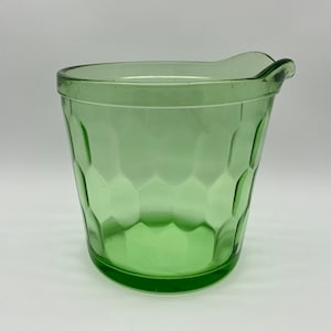 Puede incluir: Una jarra de vidrio verde con un patrón hexagonal. La jarra tiene un pico para verter fácilmente. El vidrio es de color verde claro. La jarra se utiliza probablemente para servir bebidas.