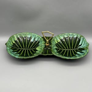 May include: A green ceramic serving dish with a leaf design. The dish has two sections and a gold-colored handle. The leaf-shaped sections have a glossy finish and are in shades of green.
