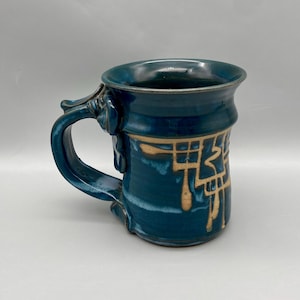 May include: A handcrafted ceramic mug in shades of teal and blue with a decorative handle. The mug features a gold-colored geometric design around the body. The mug is a unique piece of pottery.