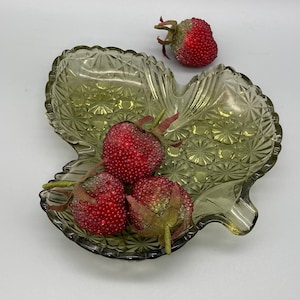 May include: A green glass leaf-shaped dish with four red strawberries inside. The dish has a raised, textured pattern.