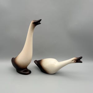 May include: Two ceramic goose figurines with a smooth, cream-colored finish and dark brown gradient shading on the base, wings, and head. One goose stands upright with its neck extended, while the other is positioned horizontally.