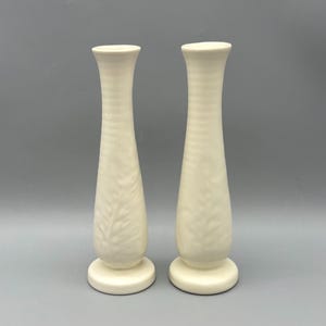 May include: A pair of tall, slender, off-white ceramic bud vases with a flared rim and a circular base. Each vase features a subtle, embossed leaf pattern on the lower body and a smooth, matte finish throughout the exterior surface.