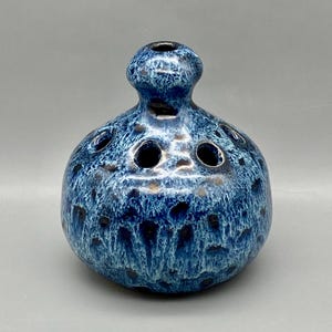 May include: A blue ceramic bud vase with a speckled glaze and a round body. The vase has a small opening at the top and several holes around the sides for holding flowers.