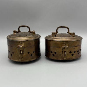 May include: Two antique brass containers with handles and decorative cutouts. The round boxes have hinged lids and clasps. The brass has a warm, aged patina. The containers are likely used for storage or display.