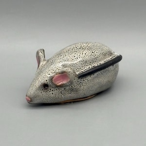 May include: A ceramic mouse figurine with a speckled gray body, black accents, and pink ears and nose. The mouse has a lid on its back and is likely a decorative trinket box.