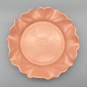 May include: A pale pink ceramic plate with a scalloped edge. The plate is round and has a smooth, glossy finish.