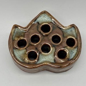 May include: A brown and white ceramic flower frog with eight holes for arranging flowers. The frog is shaped like a leaf and has a mottled glaze.