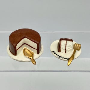 May include: A miniature chocolate cake with white frosting and a slice of cake on a plate with a gold cake server. The plate has the word "Hollywood" written on it.