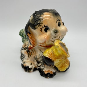 May include: A ceramic figurine of a cartoon dog with black and white fur, wearing a yellow and brown hat. The dog is holding a yellow and brown pipe in its mouth.