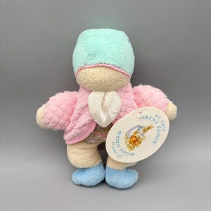 May include: A plush toy of Jeremy Fisher from Beatrix Potter's stories. The toy is light pink with a mint green cap and blue feet. It has a white tag that reads "Jeremy Fisher My First Fisher" and "Beatrix Potter on eden gift."