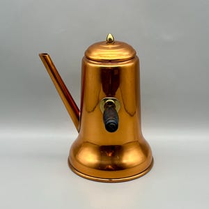 May include: A copper-colored vintage tea kettle with a long spout and a black handle. The kettle has a rounded base and a lid with a small, gold-colored finial. The overall design is sleek and classic, suitable for kitchen decor.