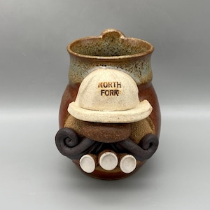 May include: A handcrafted ceramic mug with a unique design. The mug features a tan hat with the text "NORTH FORK", a brown mustache, and three white circles. The mug has a brown and tan speckled finish.