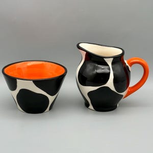 May include: A ceramic creamer pitcher and a matching bowl, both featuring a black and white cow-print pattern. The pitcher has a bright orange handle and a cream-colored interior, while the bowl has a vibrant orange interior finish.