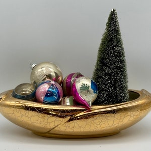 May include: A gold ceramic bowl with a crackled finish holds a small artificial Christmas tree and a collection of vintage glass ornaments in shades of pink, blue, and silver.