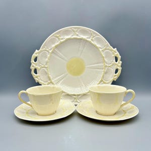 May include: A cream-colored tea set featuring two teacups with matching saucers and a serving plate. The set has a delicate, textured design with a pale yellow accent. The plate has a scalloped edge.