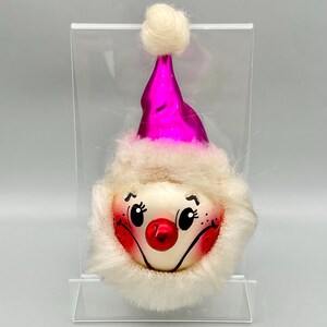 May include: A vintage Christmas ornament shaped like a smiling face. The ornament features a pink hat with a white pom-pom, a fluffy white beard, and a red nose. The face has black eyes and rosy cheeks.