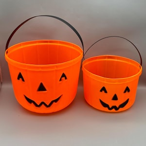 May include: Two orange plastic Halloween buckets with black handles. Each bucket has a carved jack-o'-lantern face. The larger bucket is approximately 9 inches tall, and the smaller bucket is about 6 inches tall.