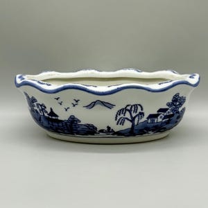 May include: White ceramic bowl with a scalloped rim and blue painted landscape design. The bowl features a detailed illustration of a village scene with trees, houses, and flying birds. A decorative piece for home decor.