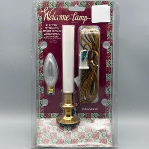 May include: An electric Welcome Lamp in original packaging with a brass-colored base, white candle-style sleeve, and a clear light bulb. The packaging features red and green checkered patterns, text detailing an auto on/off sensor, and a 6 ft. cord.