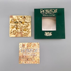 May include: A green box with gold lettering that reads "Picturesque" and "The Figurines from Nursery Kingdom." The box also has a gold logo that reads "Harmony Kingdom." The box is open and a white tile with a relief sculpture of cherubs is visible inside.