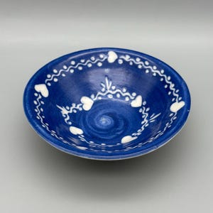 May include: A round, blue ceramic bowl with a white heart and dot design around the rim. The bowl's interior has a spiral pattern. The bowl is a decorative piece for serving or display.