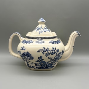 May include: Cream-colored ceramic teapot with blue floral patterns. The teapot features a curved spout, a handle, and a lid with a finial. The floral design includes flowers and leaves. The teapot is a classic design for serving tea.