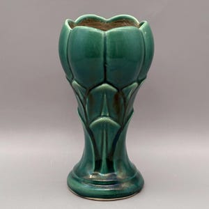 May include: A deep green ceramic vase with a glossy finish and a sculpted, petal-like exterior. The vase features a wide, scalloped rim that tapers down to a narrow neck, expanding into a circular base. The glaze shows subtle dark shading.