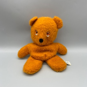 May include: An orange plush teddy bear with a black nose and orange eyes. The bear has a fuzzy texture and is sitting upright. The bear has a small tag attached to its foot.