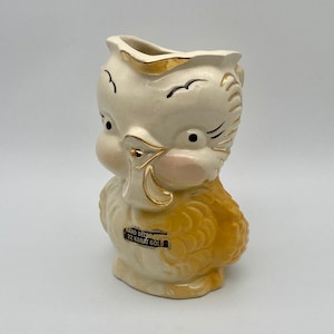 May include: A ceramic pitcher shaped like a yellow chick with hand-painted details. The chick has a pink beak and black eyes. The pitcher is decorated with 22 karat gold accents and reads "Hand Decorated 22 Karat Gold".