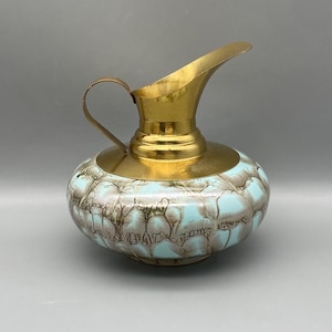May include: A vintage pitcher with a brass spout, handle, and rim. The body of the pitcher is a light blue and brown pattern. The pitcher has a rounded base and a curved spout.