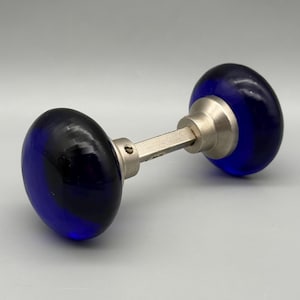 May include: A pair of blue glass door knobs with a silver-colored metal connecting rod. The glass knobs are oval-shaped and have a deep blue color. The metal rod is a rectangular shape.