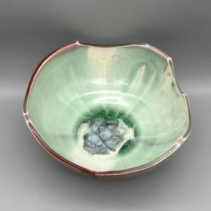 May include: A handmade ceramic bowl with a unique, organic shape. The bowl features a light green interior with a darker green center and a dark red rim. The interior has a textured, crackled glaze, and the bowl is approximately 10 inches in diameter.