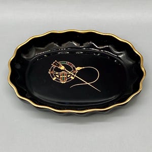 May include: Black oval trinket dish with a gold rim and a gold design of a compass and needle on the inside.