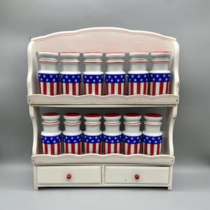 May include: White wooden spice rack with two shelves and two drawers. The rack is decorated with red trim and has ten small jars with red, white, and blue American flag designs.