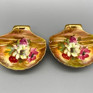 May include: Two small, gold-rimmed, shell-shaped dishes with floral designs. The dishes are painted with pink, white, and red flowers on a brown background.