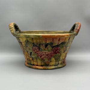 May include: A decorative ceramic basket with two handles. The basket features a woven design with embossed red roses and green leaves. The color palette includes shades of green, brown, and red, creating a rustic aesthetic.