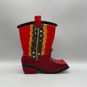 May include: A decorative wooden cowboy boot in shades of red, brown, black, green, and gold. The boot features a detailed design with cut-out accents and a classic western style. The boot is approximately 10 inches tall.