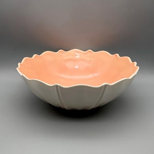May include: A large, decorative bowl with a white exterior and a peach-colored interior. The bowl has a scalloped rim and a fluted design on the exterior. The bowl is likely made of ceramic or porcelain and is suitable for serving or display.