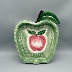 May include: An apple-shaped ceramic dish featuring a textured green sponge-painted border and a dark brown stem with a green leaf. The center of the dish displays a hand-painted illustration of a red apple with two green leaves on a white background.
