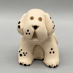 May include: White ceramic dog figurine with black spots. The dog is sitting with its head tilted up and has a playful expression. The figurine has a textured surface and is hand-painted.