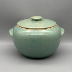 May include: A round, teal-colored ceramic casserole dish with a matching lid. The dish has two small handles on either side and a small knob on the lid. The dish is glazed with a glossy finish.