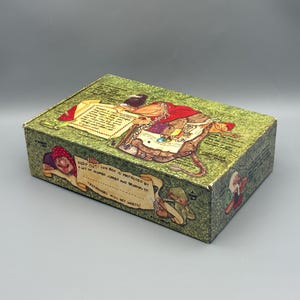 May include: A rectangular cardboard box with a green textured background featuring whimsical illustrations of fantasy creatures. The front panel displays the text: KEEP OUT! THIS BOX IS PROTECTED BY A LOT OF MUMBO JUMBO AND BELONGS TO: TRESPASSERS WILL GET WARTS!