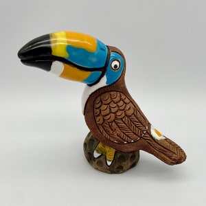 May include: A ceramic toucan figurine with a yellow, blue, and black beak, brown body with detailed feather markings, and white accents. The toucan is perched on a brown base with white spots.