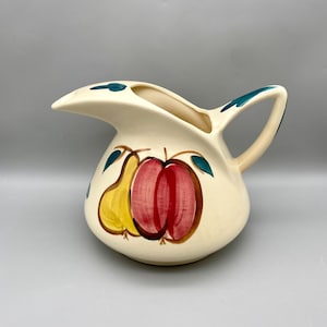 May include: A white ceramic pitcher with a green handle and a pear and apple design. The pitcher has a wide spout and a rounded body.