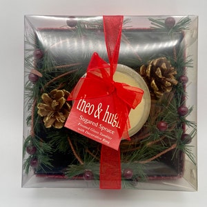 May include: A clear plastic box with a red ribbon and bow contains a frosted glass candle holder with a white candle. The candle holder is surrounded by a green pine wreath with pine cones and red berries. The label on the box reads "theo & hugh Sugared Spruce Frosted Glass Tumbler with Decorative Ring."