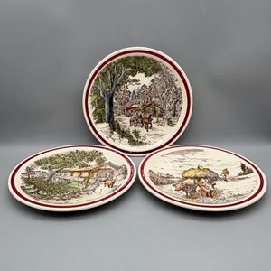 May include: Three ceramic plates with a rural landscape design. The plates have a white background with a detailed illustration in shades of brown, green, and red. Each plate features a different scene, with a red border.