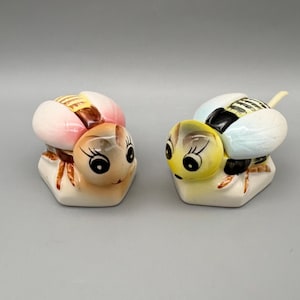 May include: Two ceramic bee figurines. One bee has pink and brown wings, the other has yellow and blue wings. Both have black and white eyes and are on a white base.