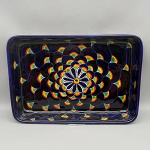 May include: Rectangular ceramic serving dish with a dark blue background and a floral design in the center. The floral design features a white flower with a yellow center, surrounded by orange, green, and yellow accents.